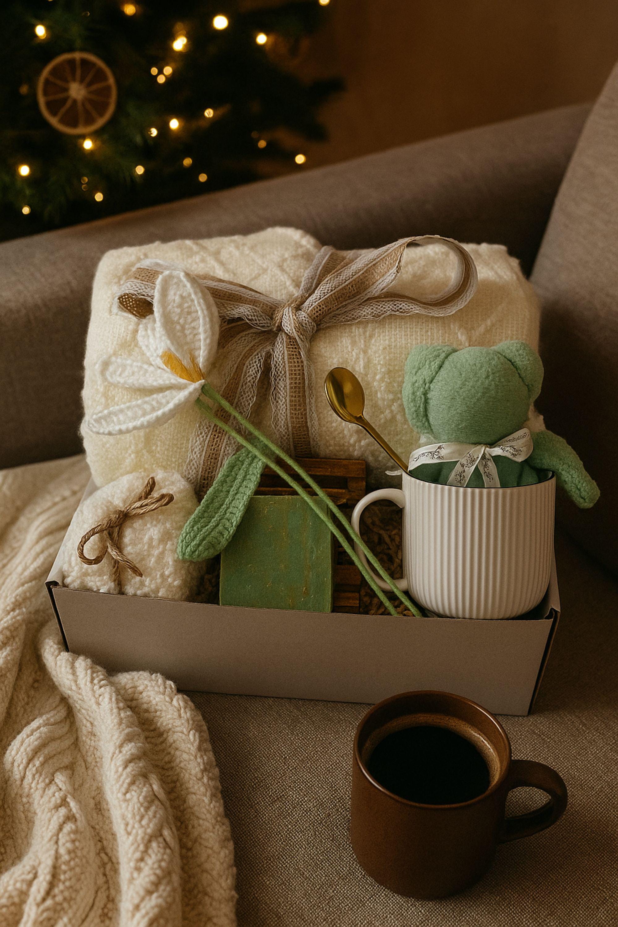Christmas cozy spa gift box with white blanket tied in lace ribbon, crochet tulip, white ceramic mug with crochet bear, handmade green soap on bamboo tray, gold spoon, and spa puff arranged in a gift box with crinkle paper. Holiday comfort and relaxation gift for women.