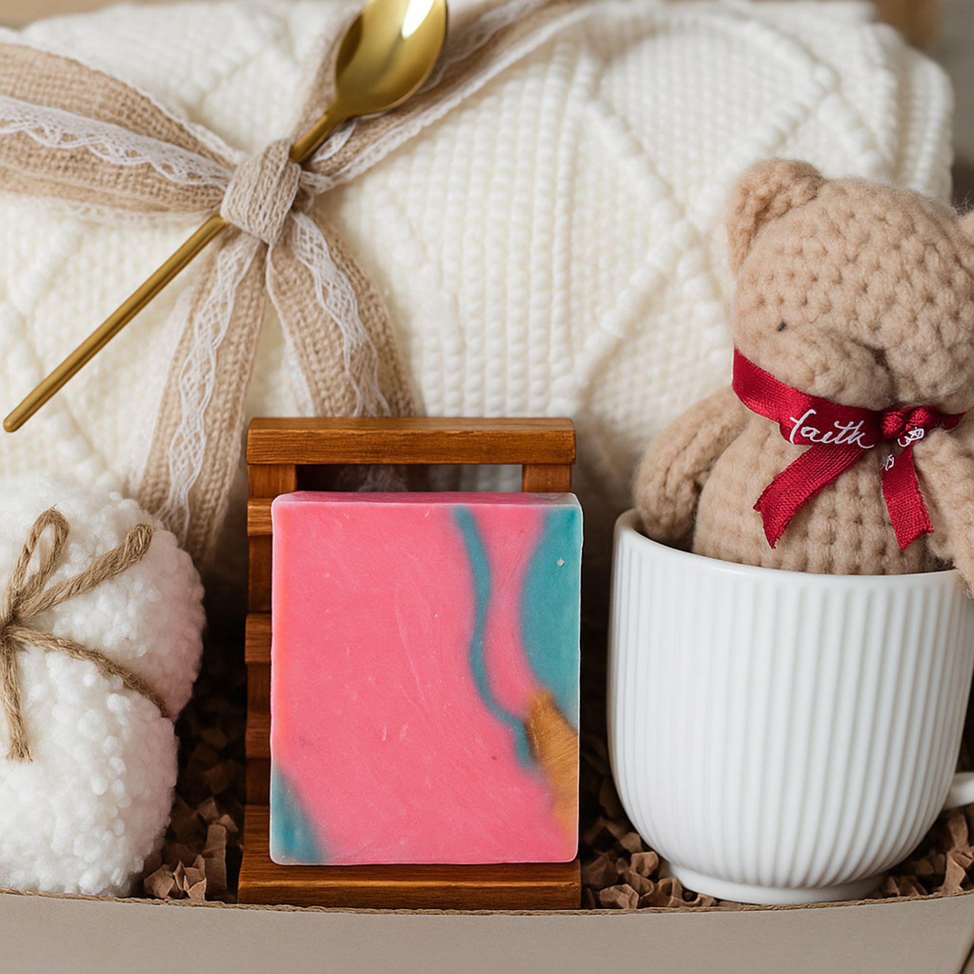 Winter self-care gift box featuring a cozy white blanket with ribbon, a teddy-bear-shaped washcloth, handmade pink and blue soap on a wooden tray, and a soft crocheted bear sitting in a white mug. Displayed on a rustic wood surface with snowy accents, this cozy winter spa set makes a thoughtful holiday comfort gift, pamper box, relaxation present, or get-well care package for women.