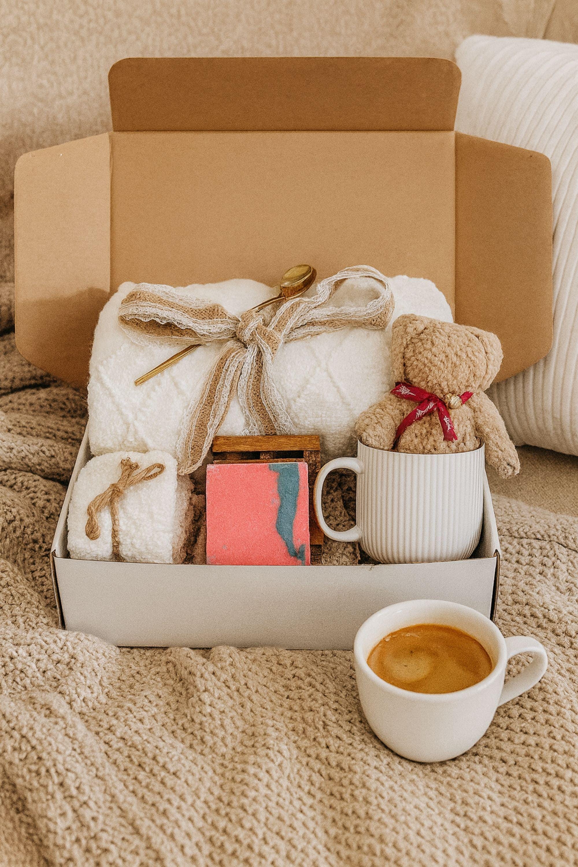 Winter self-care gift box featuring a cozy white blanket with ribbon, a teddy-bear-shaped washcloth, handmade pink and blue soap on a wooden tray, and a soft crocheted bear sitting in a white mug. Displayed on a rustic wood surface with snowy accents, this cozy winter spa set makes a thoughtful holiday comfort gift, pamper box, relaxation present, or get-well care package for women.