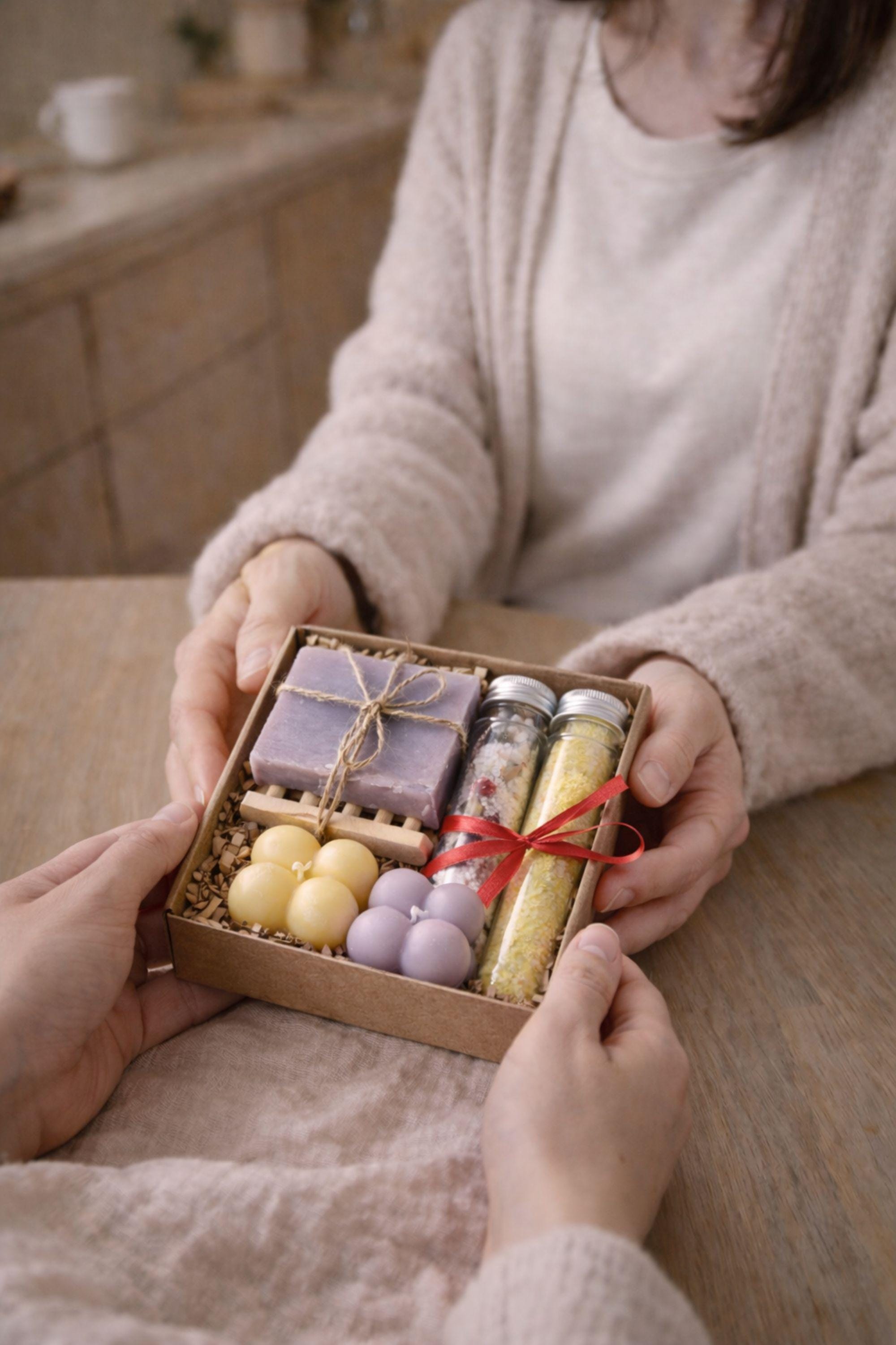 Lifestyle photo showing a small 6x6 inch handmade gift box for mom held in hands to demonstrate real size and scale. Open kraft box reveals colorful bubble candles, natural soap, and bath salts, helping buyers understand dimensions and presentation of this cozy, thoughtful gift for mom.