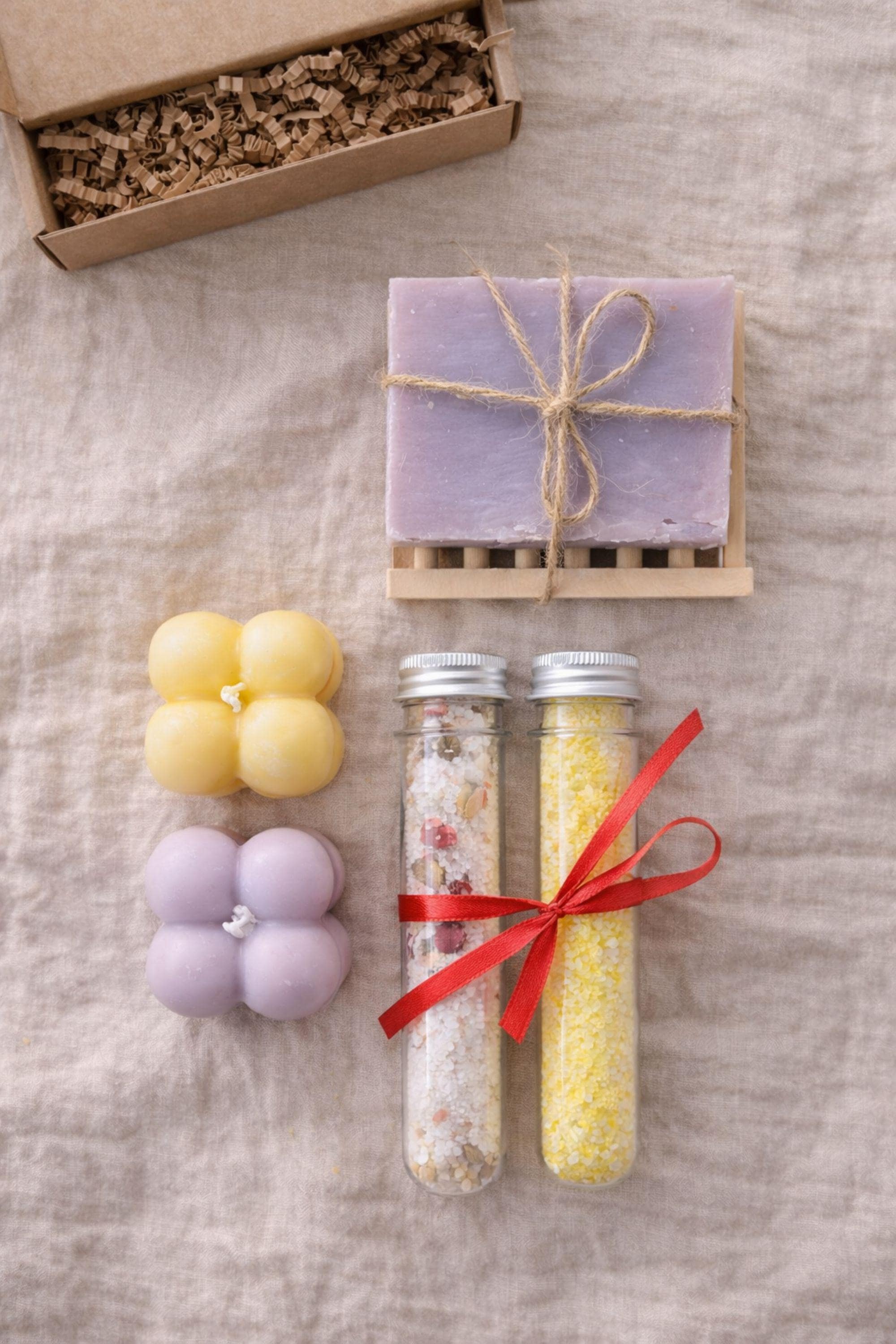 Flat lay photograph showing everything included in a gift for mom box: handmade bubble candles, natural soap on a wooden soap dish, and two glass tubes filled with bath salts. Items are neatly arranged to clearly display contents of a small 6x6 inch handmade gift box designed for thoughtful gifting.