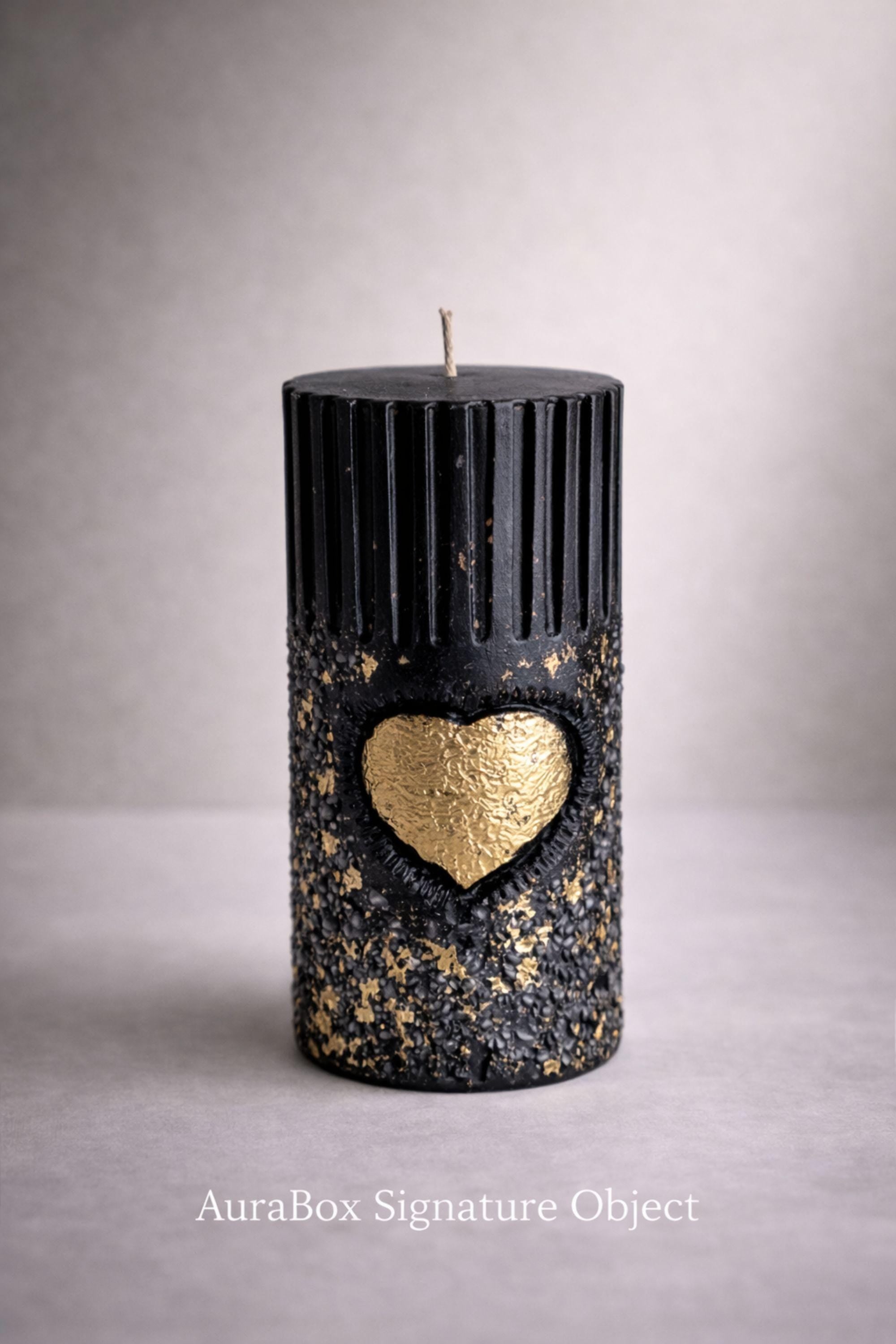 Black sculptural pillar candle made of deep black wax with a ribbed cylindrical form and textured lower section. A hand-applied gold heart accent is centered on the front, surrounded by subtle gold flecks. Photographed front-facing on a neutral background to emphasize form, proportions, and object presence. Designed as a symbolic wax object with candle function as secondary use.