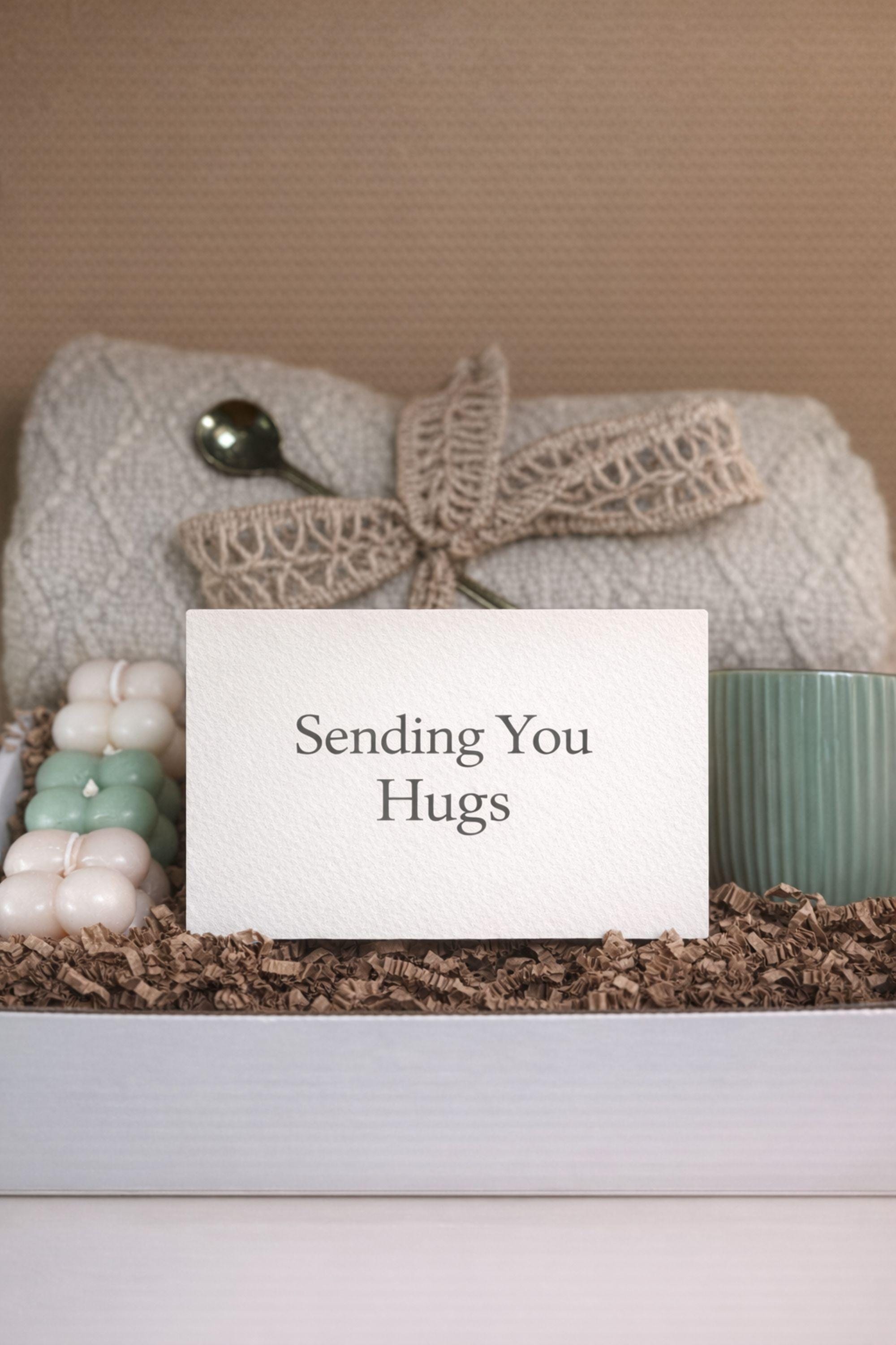 Sending You Hugs grief support gift box with sage knit blanket, ribbed green ceramic mug, handmade two-tone artisan soap, stainless steel spoon, and three bubble candles arranged inside kraft box on warm beige linen background. Sympathy care package designed for comfort during difficult times, loss support, thinking of you gift, and emotional encouragement.