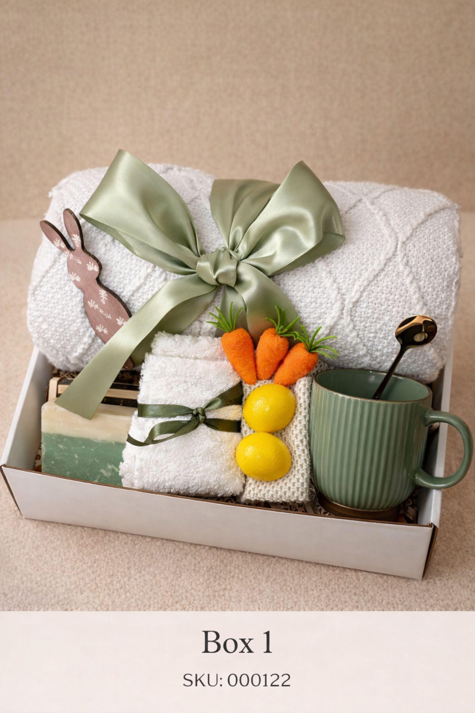 Easter gift box for women featuring a cozy knit blanket tied with green ribbon, ceramic mug with spoon, handmade artisan soap, soft socks, two lemon shaped soaps, decorative spring carrots and bunny accent arranged inside a gift box. Spring self care gift basket and Easter care package alternative to traditional Easter baskets, perfect Easter present for mom, sister, friend or women who enjoy cozy comfort gifts.