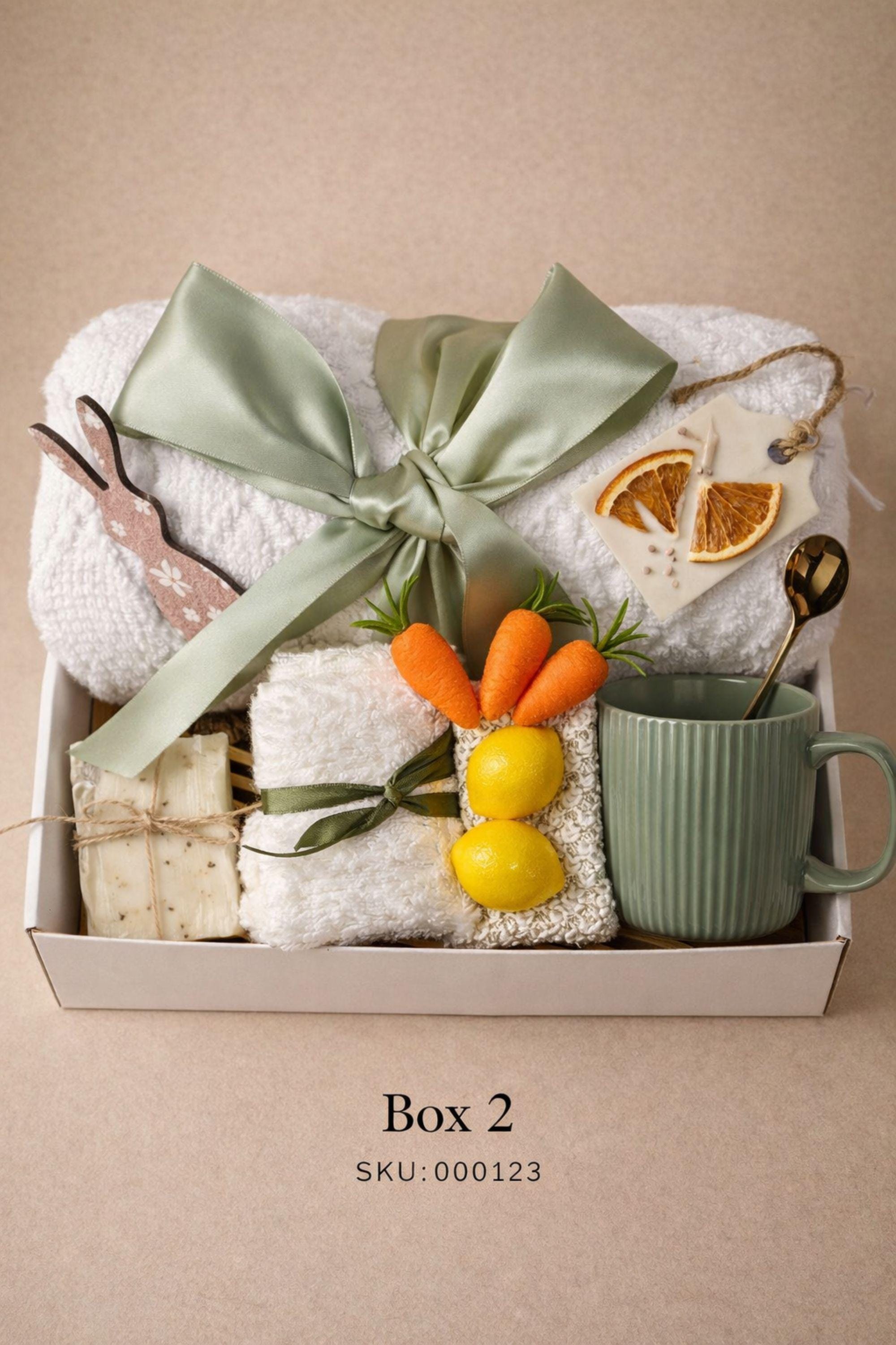 Easter gift box for women featuring a cozy knit blanket tied with green ribbon, ceramic mug with spoon, handmade artisan soap, soft socks, two lemon shaped soaps, decorative spring carrots and bunny accent arranged inside a gift box. Spring self care gift basket and Easter care package alternative to traditional Easter baskets, perfect Easter present for mom, sister, friend or women who enjoy cozy comfort gifts.