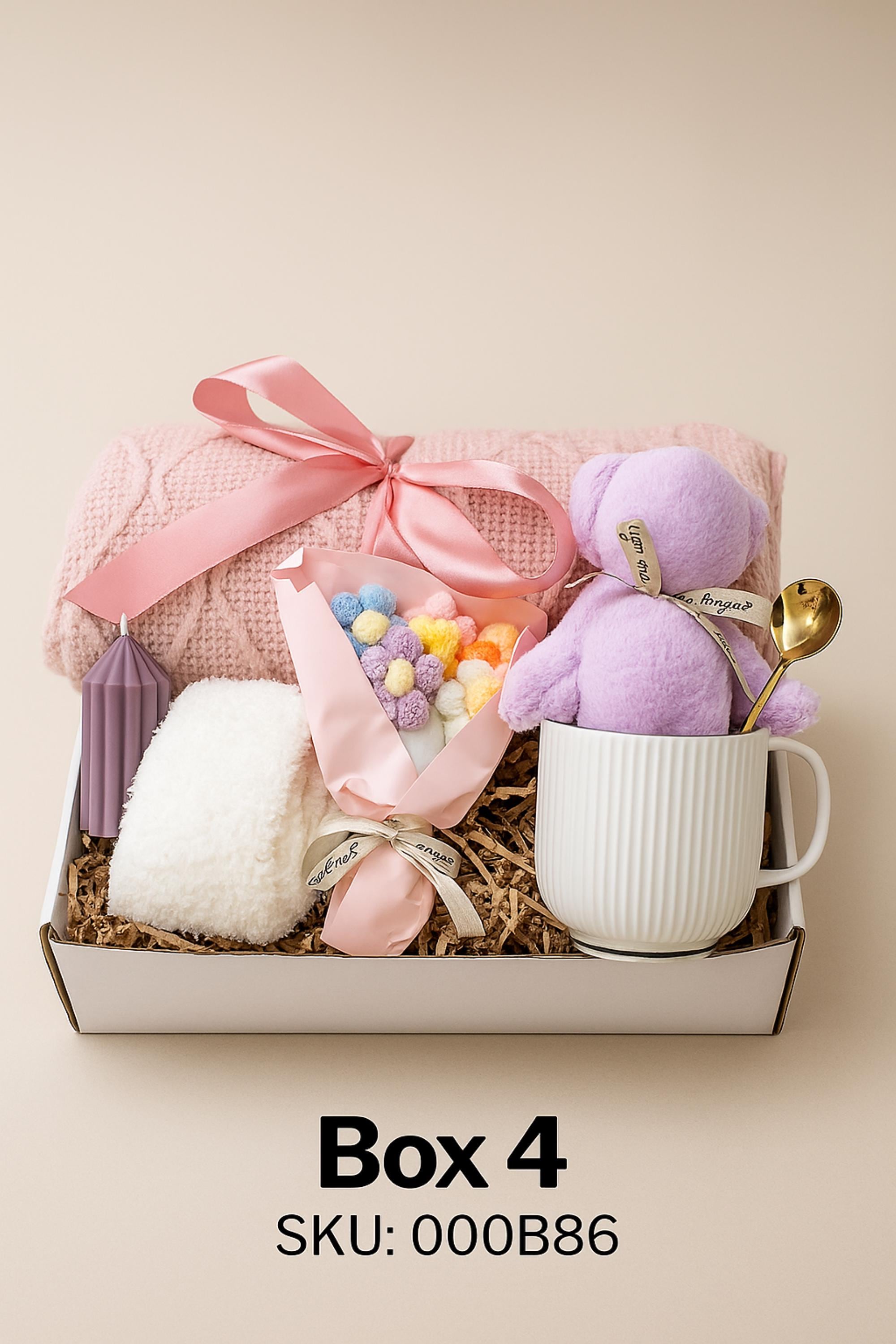 Christmas cozy spa gift box with white blanket tied in lace ribbon, crochet tulip, white ceramic mug with crochet bear, handmade green soap on bamboo tray, gold spoon, and spa puff arranged in a gift box with crinkle paper. Holiday comfort and relaxation gift for women.