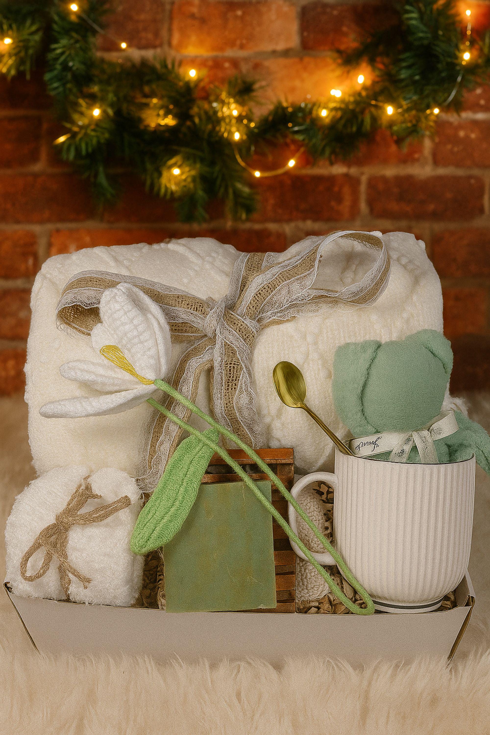 Christmas cozy spa gift box with white blanket tied in lace ribbon, crochet tulip, white ceramic mug with crochet bear, handmade green soap on bamboo tray, gold spoon, and spa puff arranged in a gift box with crinkle paper. Holiday comfort and relaxation gift for women.
