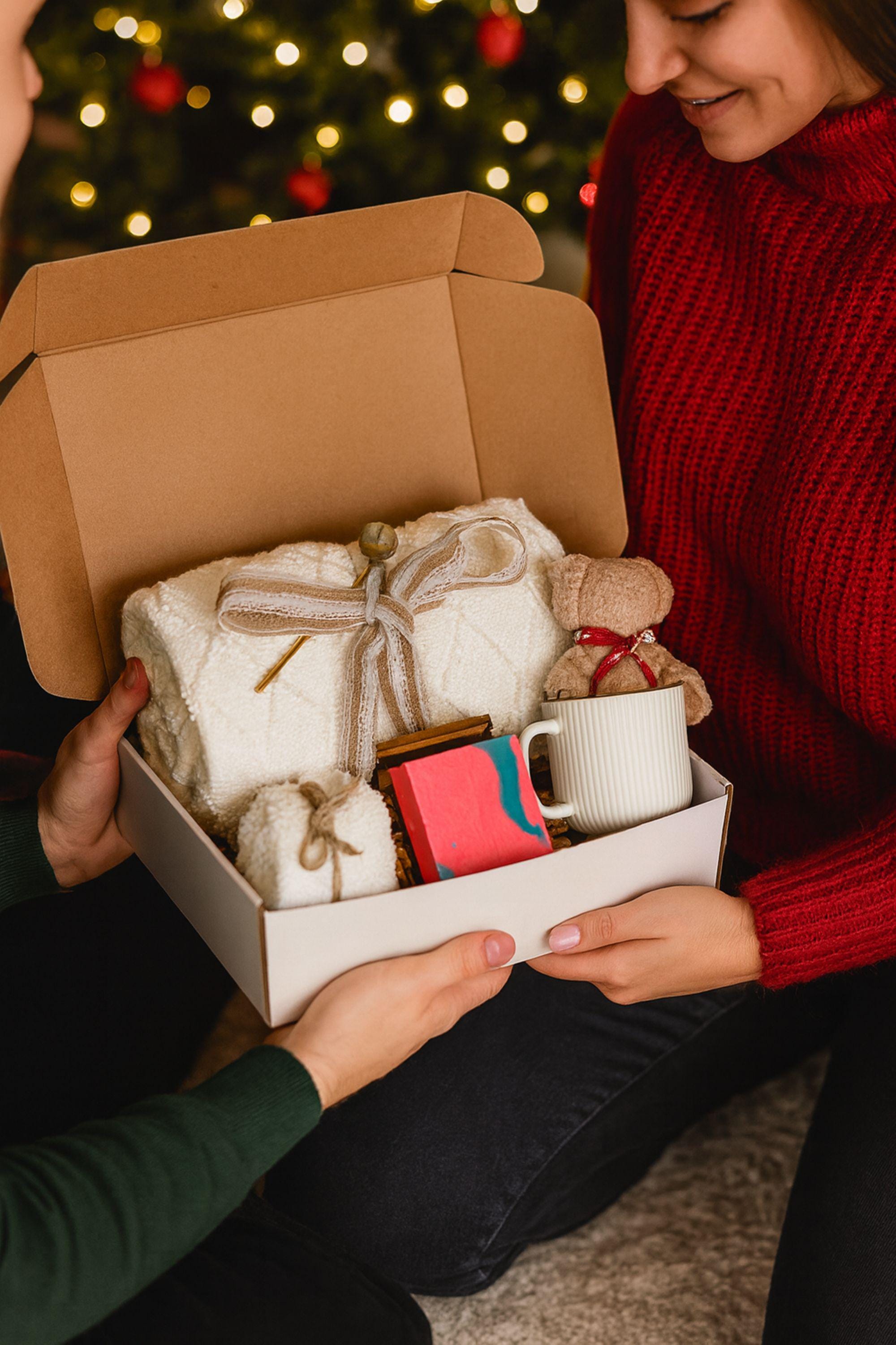 Winter self-care gift box featuring a cozy white blanket with ribbon, a teddy-bear-shaped washcloth, handmade pink and blue soap on a wooden tray, and a soft crocheted bear sitting in a white mug. Displayed on a rustic wood surface with snowy accents, this cozy winter spa set makes a thoughtful holiday comfort gift, pamper box, relaxation present, or get-well care package for women.