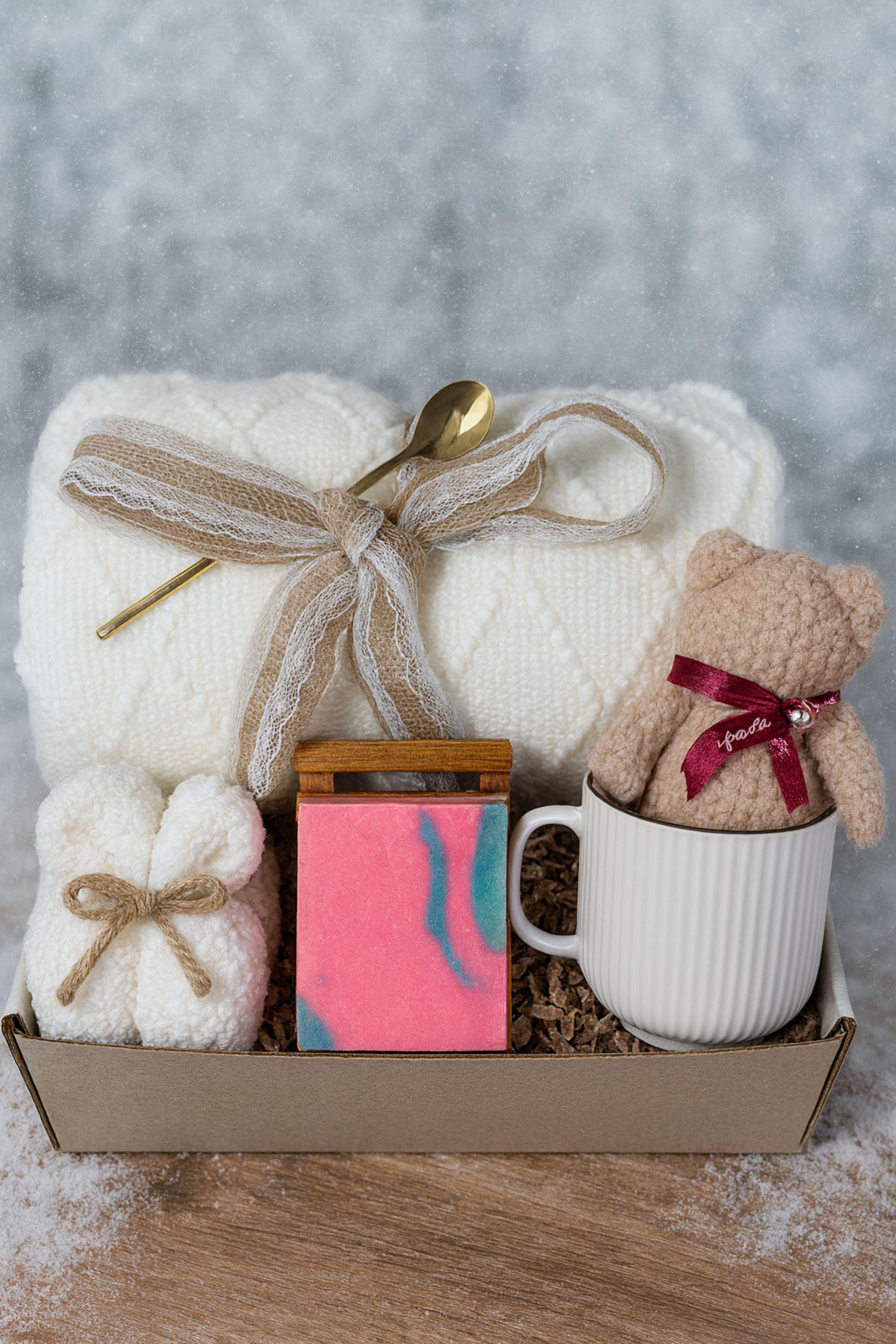 Winter self-care gift box featuring a cozy white blanket with ribbon, a teddy-bear-shaped washcloth, handmade pink and blue soap on a wooden tray, and a soft crocheted bear sitting in a white mug. Displayed on a rustic wood surface with snowy accents, this cozy winter spa set makes a thoughtful holiday comfort gift, pamper box, relaxation present, or get-well care package for women.