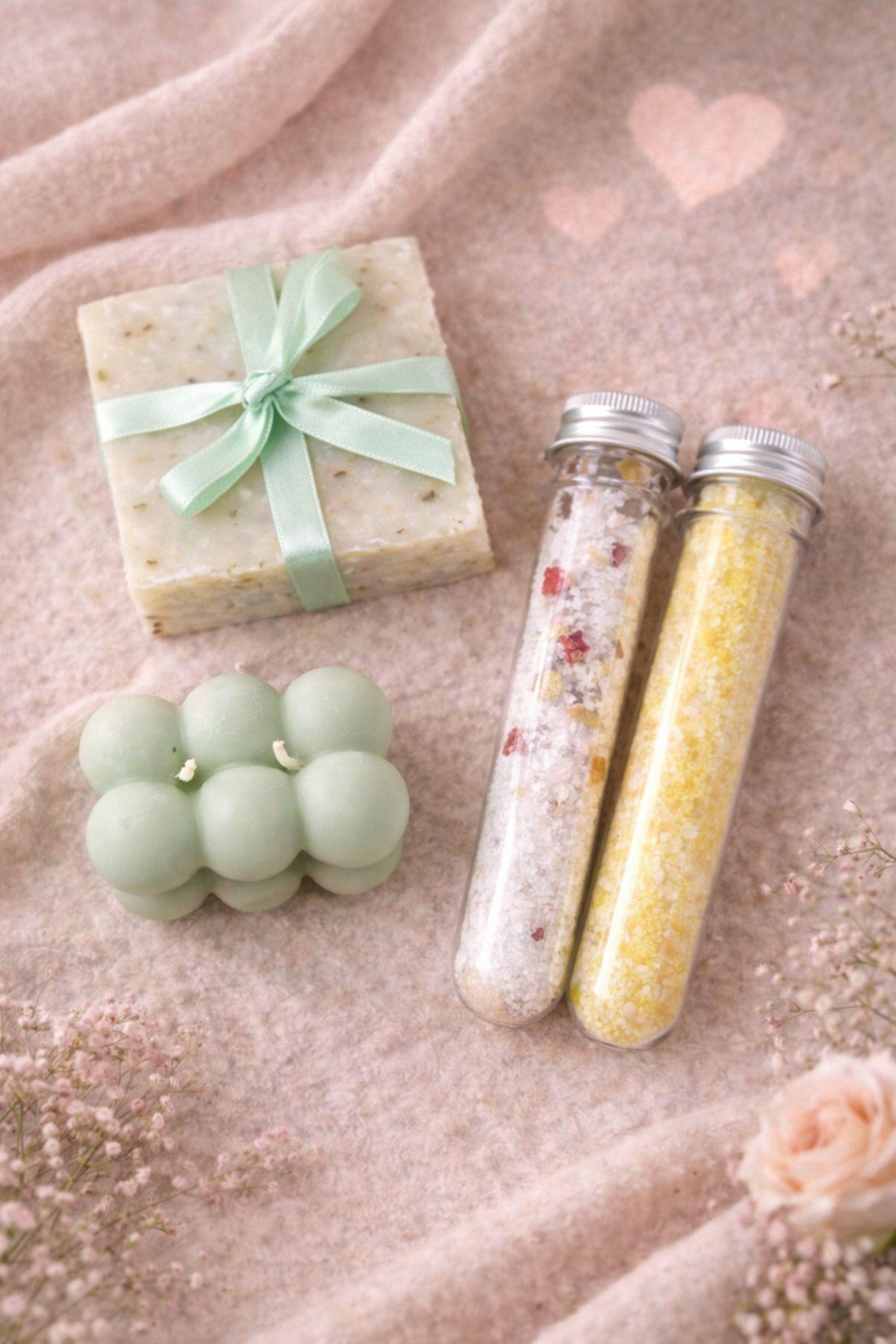Flat lay of Valentine gift box contents including sage bubble candles, handmade soap, and two glass tubes of bath salts arranged on a neutral background. Calm Valentine care package showcasing handmade items designed for comfort and relaxation, perfect as a cozy Valentine gift for her or a soft, meaningful handmade present.