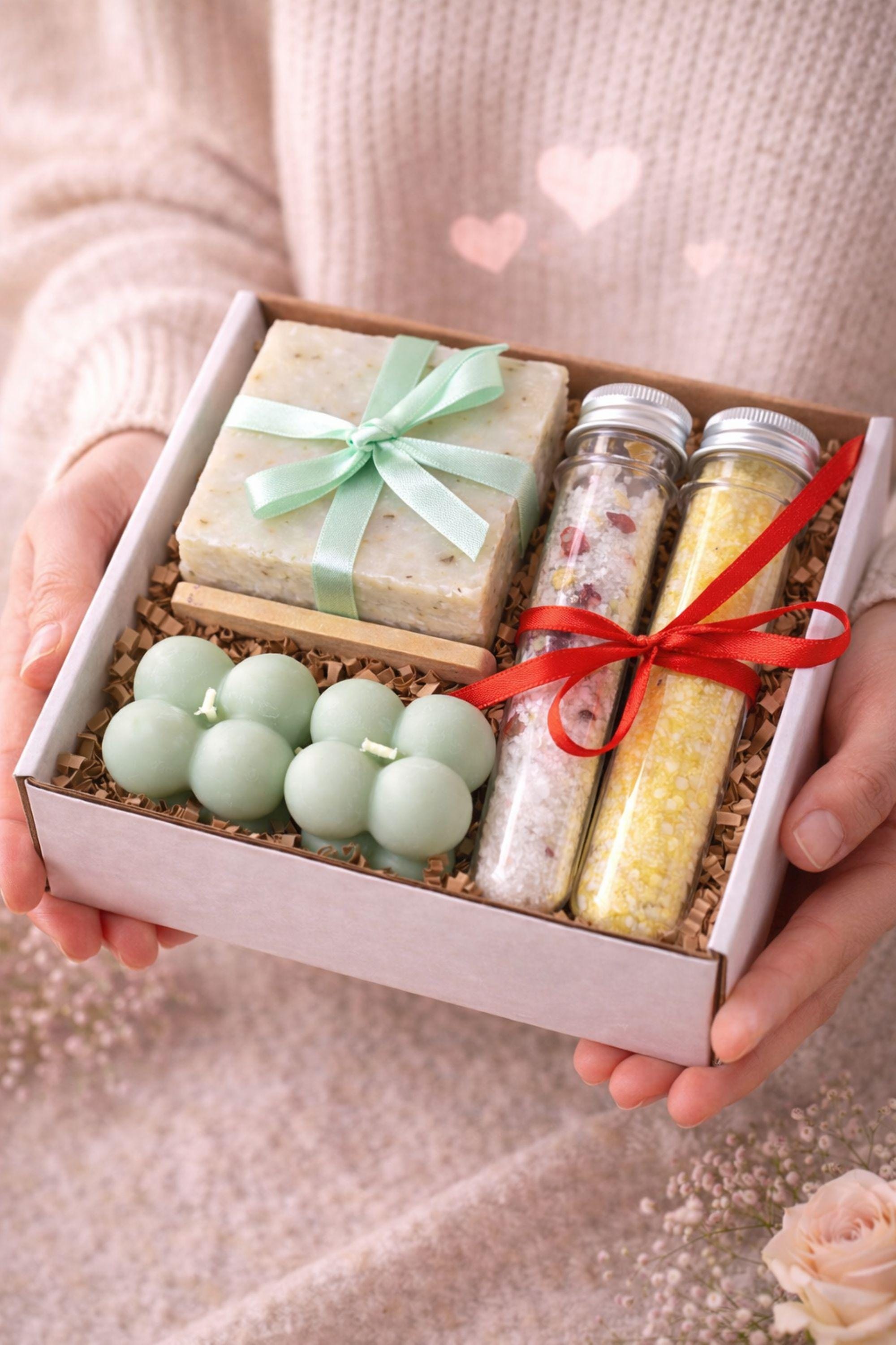 Hands holding an open Valentine gift box with sage bubble candles, natural soap, and bath salts, styled in soft neutral tones. Calm Valentine care package designed to feel gentle and comforting, ideal as a thoughtful Valentine gift for her, a cozy care box, or a handmade comfort present sent with care and intention.