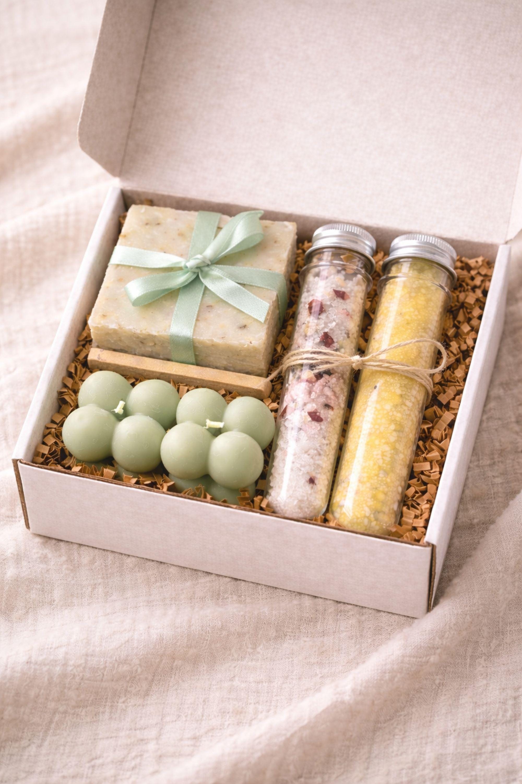 Minimalist cozy gift box displayed open on a neutral linen surface, featuring handmade sage bubble candles, natural soap tied with a soft ribbon, and two glass tubes of bath salts arranged neatly inside a white box. Calm home gift set designed for everyday self care, slow living routines, and cozy home moments, ideal as a minimalist handmade candle and soap set for a peaceful interior atmosphere.