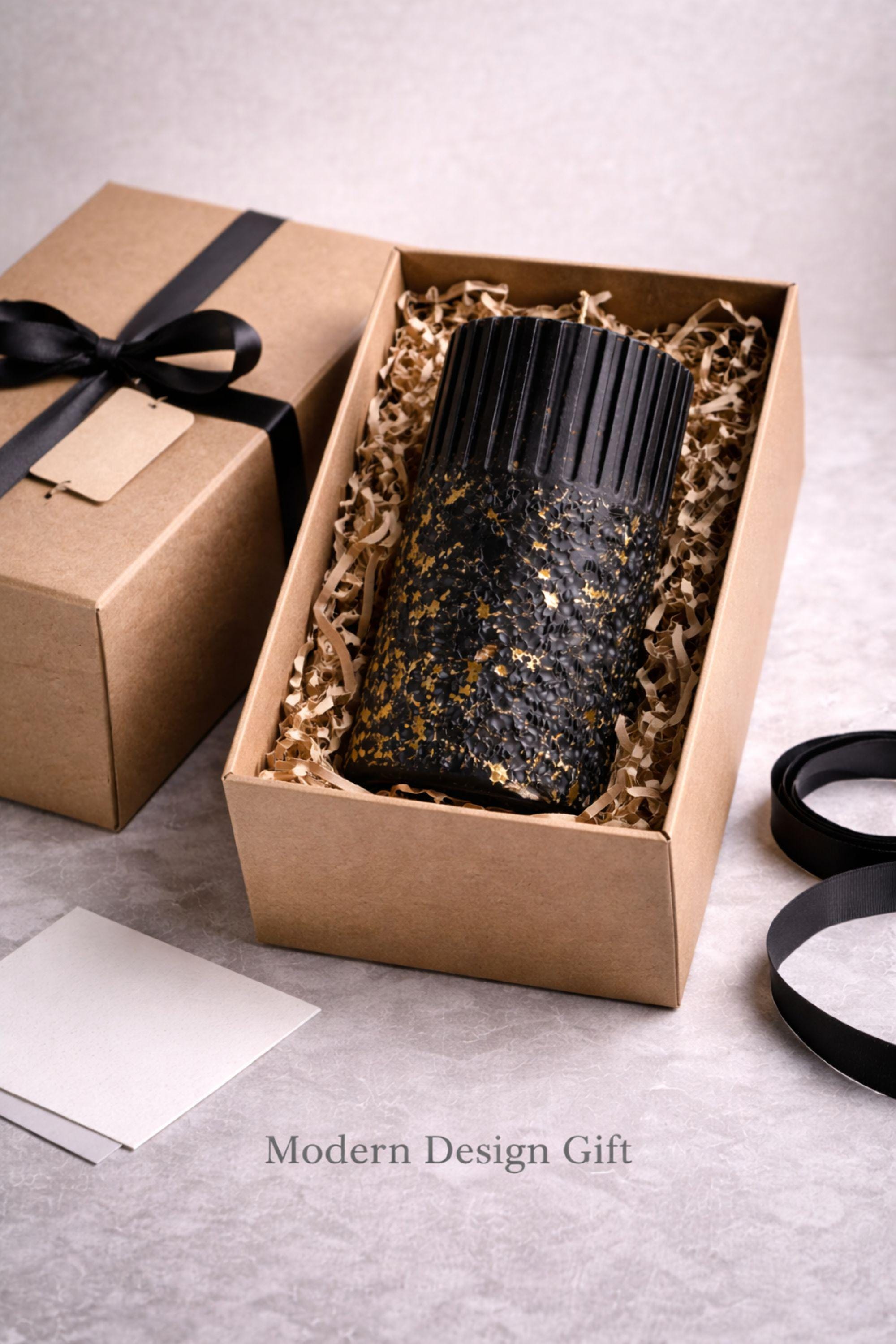 Handmade black pillar object presented in minimal gift packaging with natural filler. The sculptural wax form and gold flecks are shown ready to gift, emphasizing suitability as a neutral modern design gift without symbolic messaging.