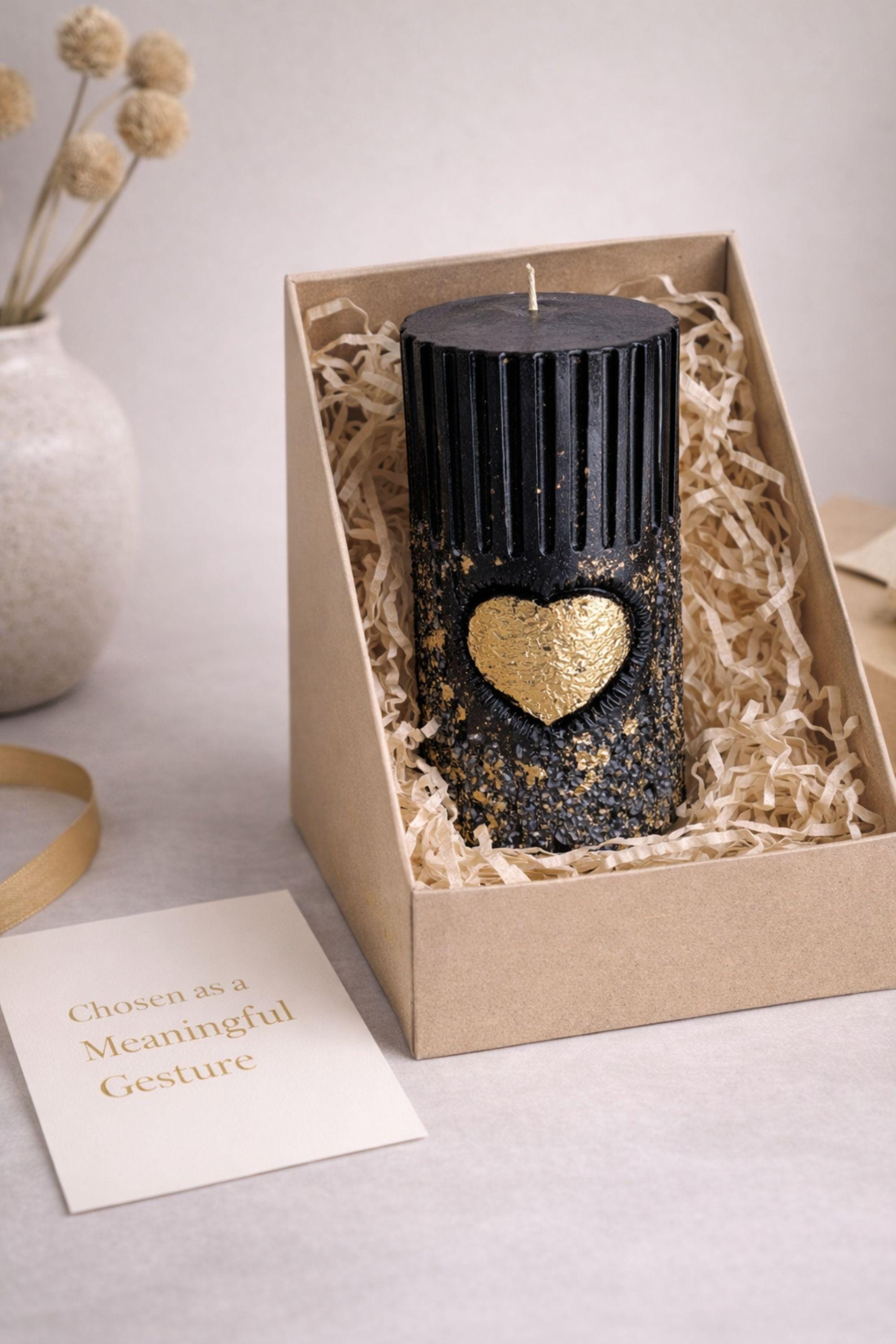 Handmade black pillar candle presented in minimal gift packaging with natural filler and a neutral card reading “Chosen as a Meaningful Gesture.” The sculptural wax object with gold heart accent is shown ready to gift, highlighting intentional presentation and suitability as a symbolic design gift.