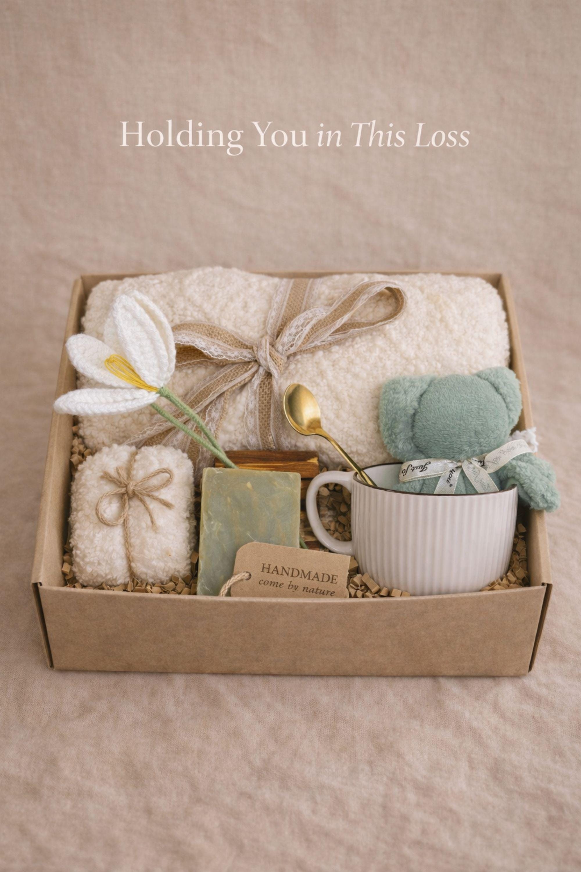 Miscarriage gift box featuring a soft cream knit blanket with lace ribbon, sage teddy bear in white ceramic mug with gold spoon, handmade soap bar, crocheted flower, and wrapped towel arranged in kraft box on neutral beige background with overlay “Holding you in This Loss.” Pregnancy loss care package designed as baby loss sympathy gift and comfort support for grieving mothers.