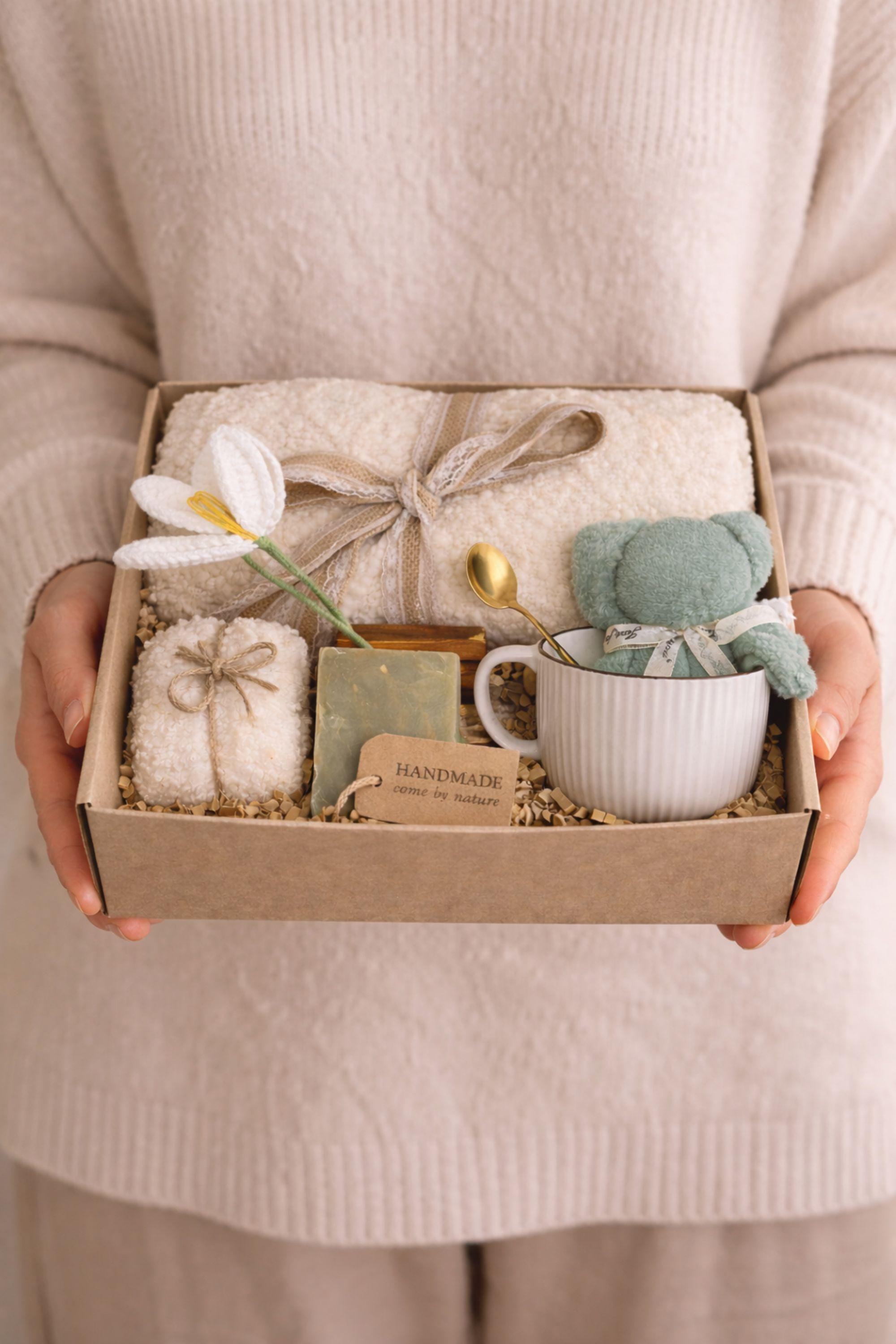 Miscarriage gift box featuring a soft cream knit blanket with lace ribbon, sage teddy bear in white ceramic mug with gold spoon, handmade soap bar, crocheted flower, and wrapped towel arranged in kraft box on neutral beige background with overlay “Holding you in This Loss.” Pregnancy loss care package designed as baby loss sympathy gift and comfort support for grieving mothers.
