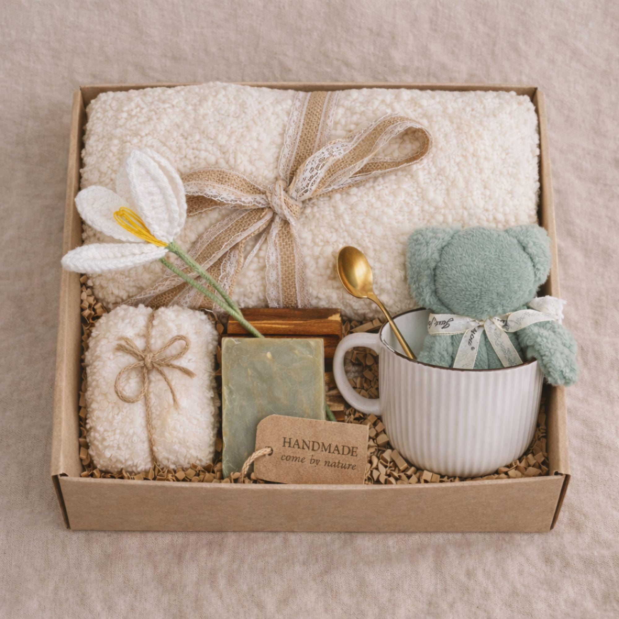 Miscarriage gift box featuring a soft cream knit blanket with lace ribbon, sage teddy bear in white ceramic mug with gold spoon, handmade soap bar, crocheted flower, and wrapped towel arranged in kraft box on neutral beige background with overlay “Holding you in This Loss.” Pregnancy loss care package designed as baby loss sympathy gift and comfort support for grieving mothers.