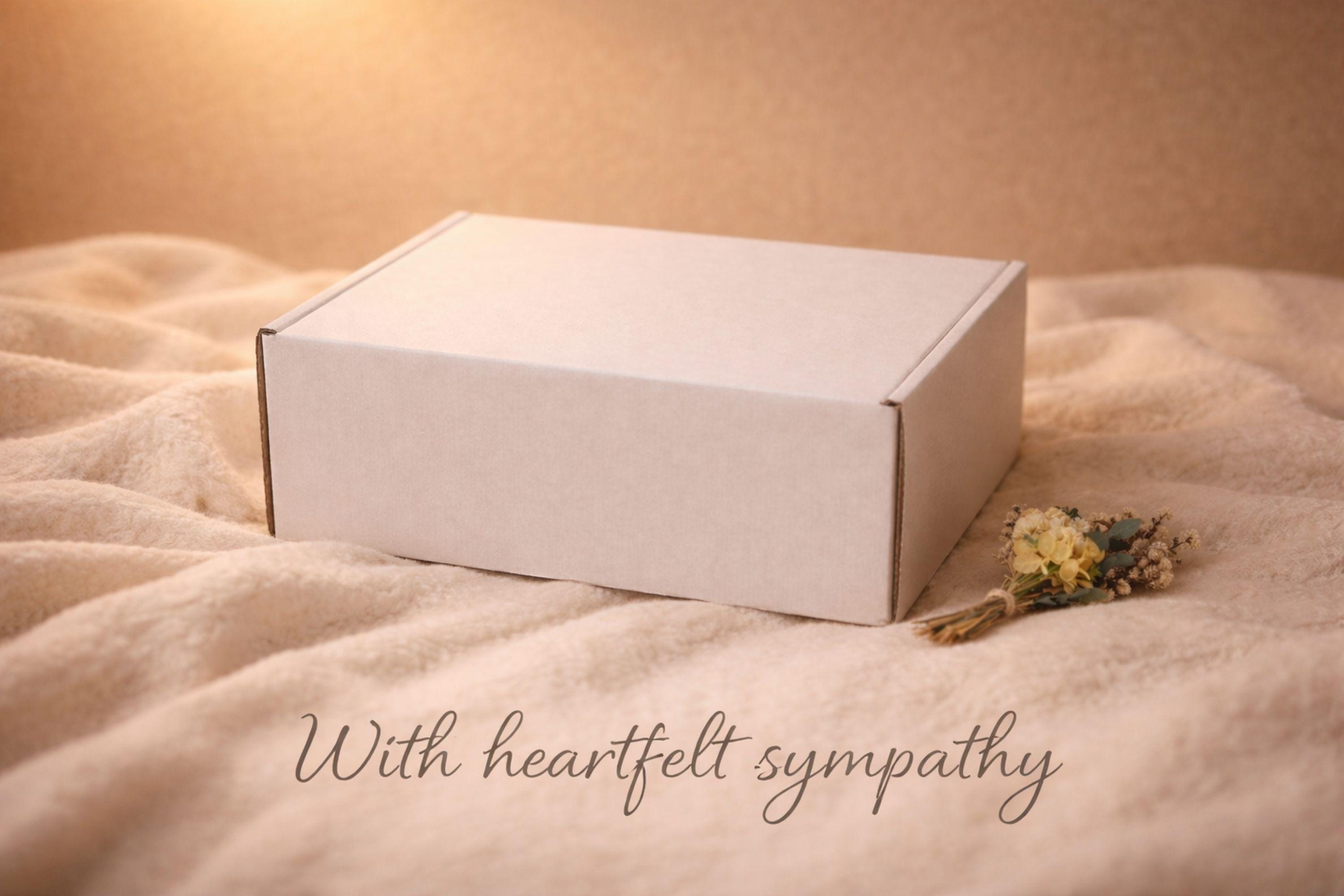 Open white sympathy gift box styled on a soft beige blanket with warm candlelight in the background. Inside the box are cozy patterned knit socks, a handmade natural soap bar tied with twine, a wooden soap holder, a mesh bath salt bag filled with visible salt crystals and botanicals, dried flowers, and a brown dog-shaped candle facing left. Text overlay reads “Thinking of you – Sending comforting care during this difficult time.”