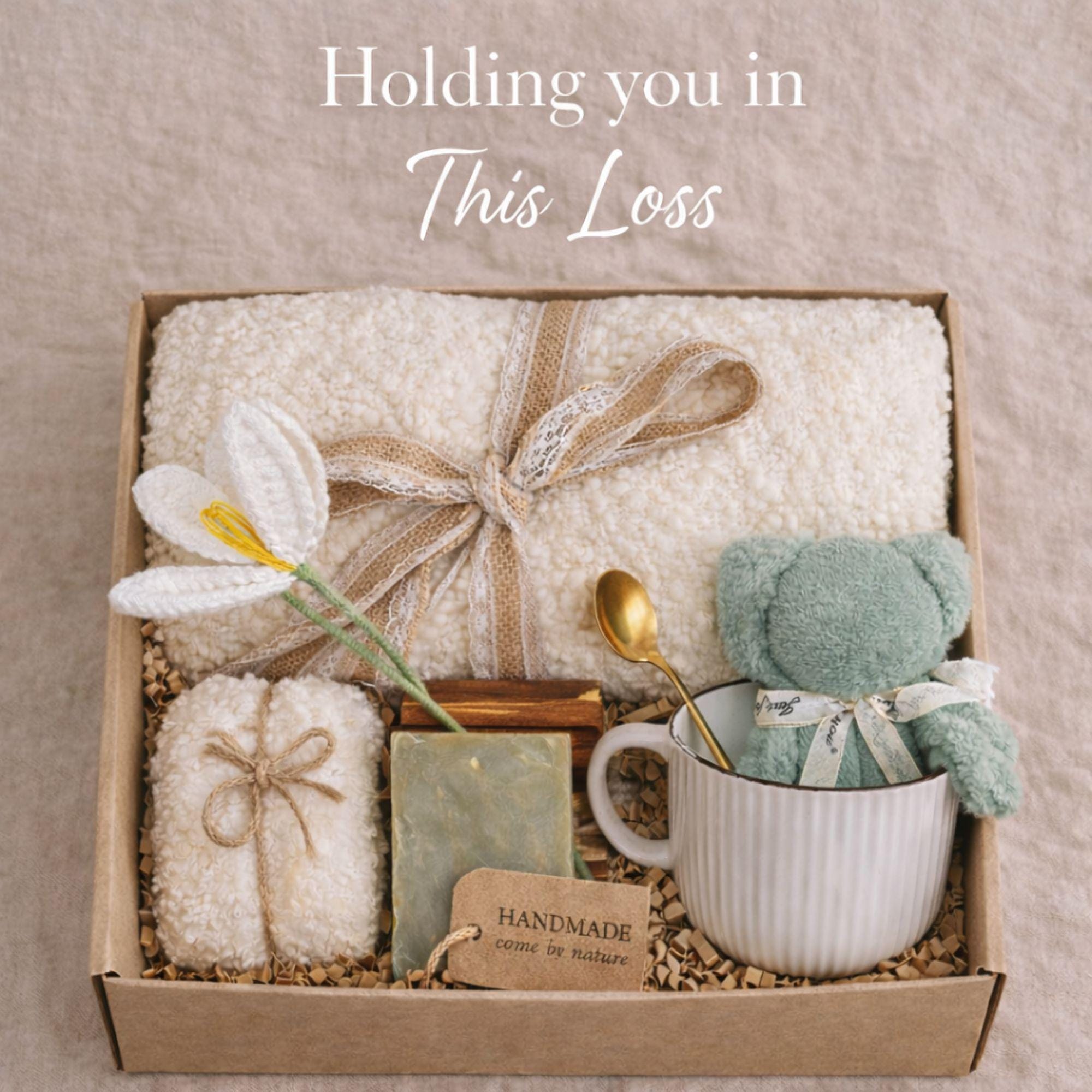 Miscarriage gift box featuring a soft cream knit blanket with lace ribbon, sage teddy bear in white ceramic mug with gold spoon, handmade soap bar, crocheted flower, and wrapped towel arranged in kraft box on neutral beige background with overlay “Holding you in This Loss.” Pregnancy loss care package designed as baby loss sympathy gift and comfort support for grieving mothers.