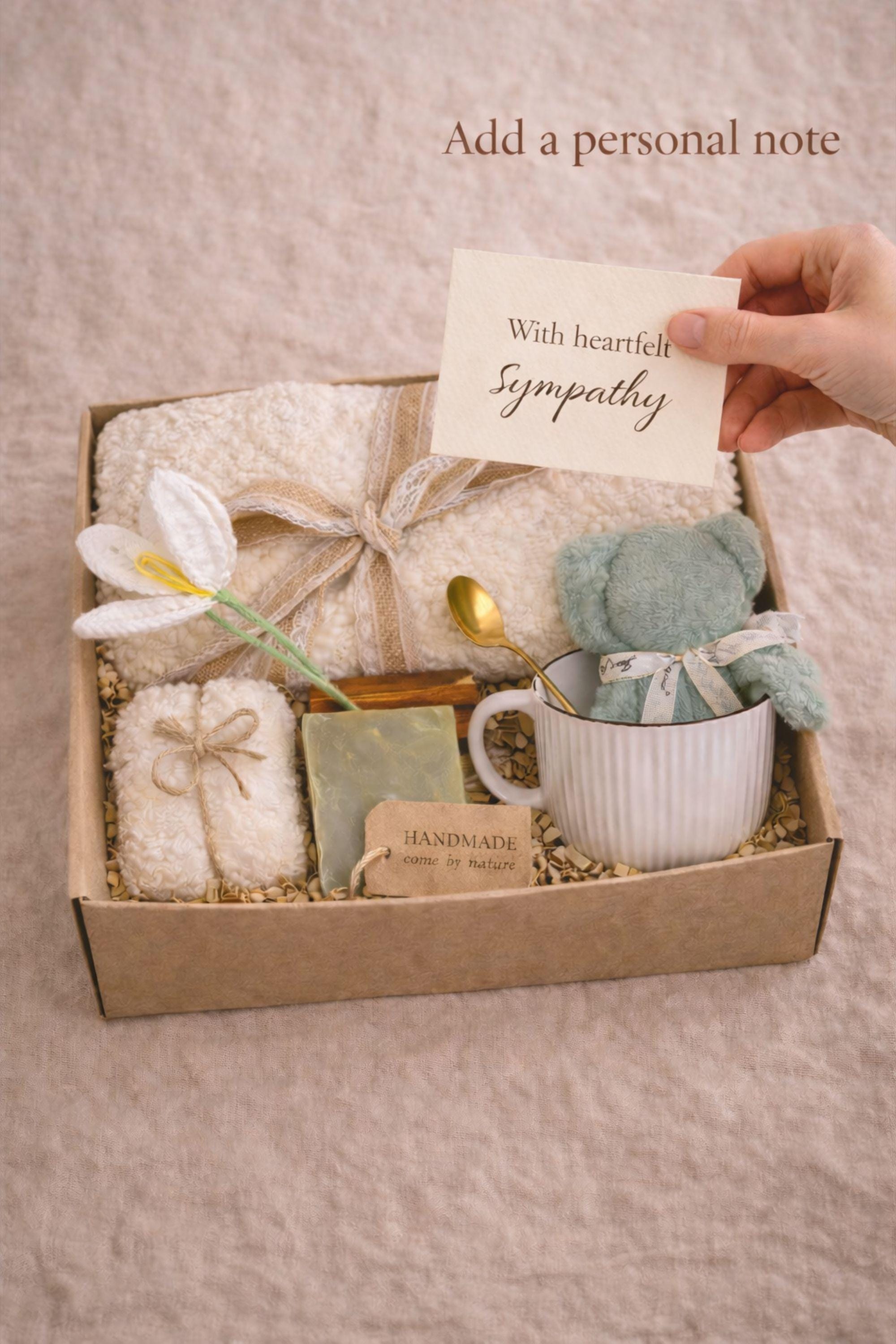 Miscarriage gift box featuring a soft cream knit blanket with lace ribbon, sage teddy bear in white ceramic mug with gold spoon, handmade soap bar, crocheted flower, and wrapped towel arranged in kraft box on neutral beige background with overlay “Holding you in This Loss.” Pregnancy loss care package designed as baby loss sympathy gift and comfort support for grieving mothers.
