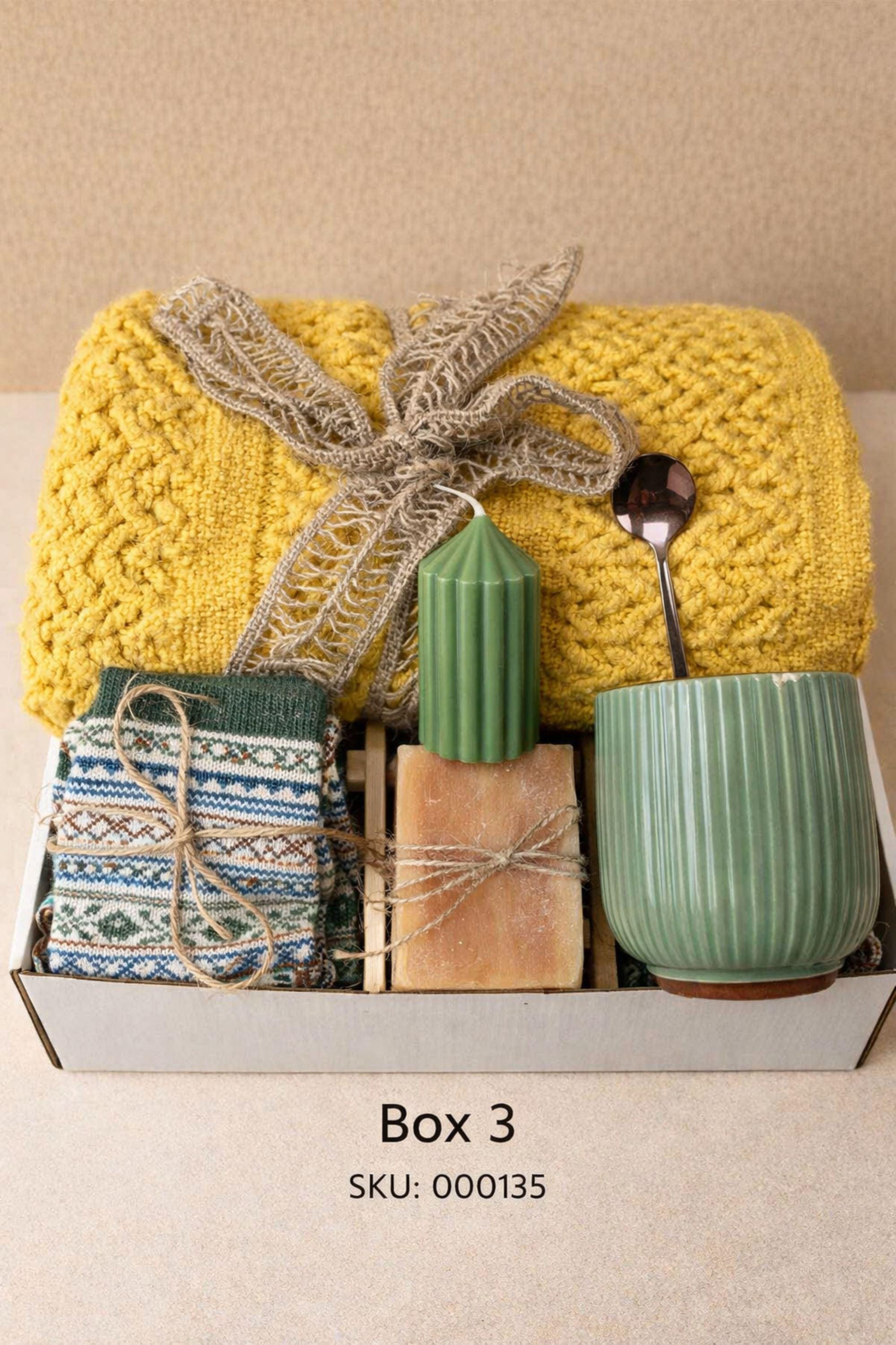 Easter gift box for women featuring a cozy knit blanket tied with green ribbon, ceramic mug with spoon, handmade artisan soap, soft socks, two lemon shaped soaps, decorative spring carrots and bunny accent arranged inside a gift box. Spring self care gift basket and Easter care package alternative to traditional Easter baskets, perfect Easter present for mom, sister, friend or women who enjoy cozy comfort gifts.