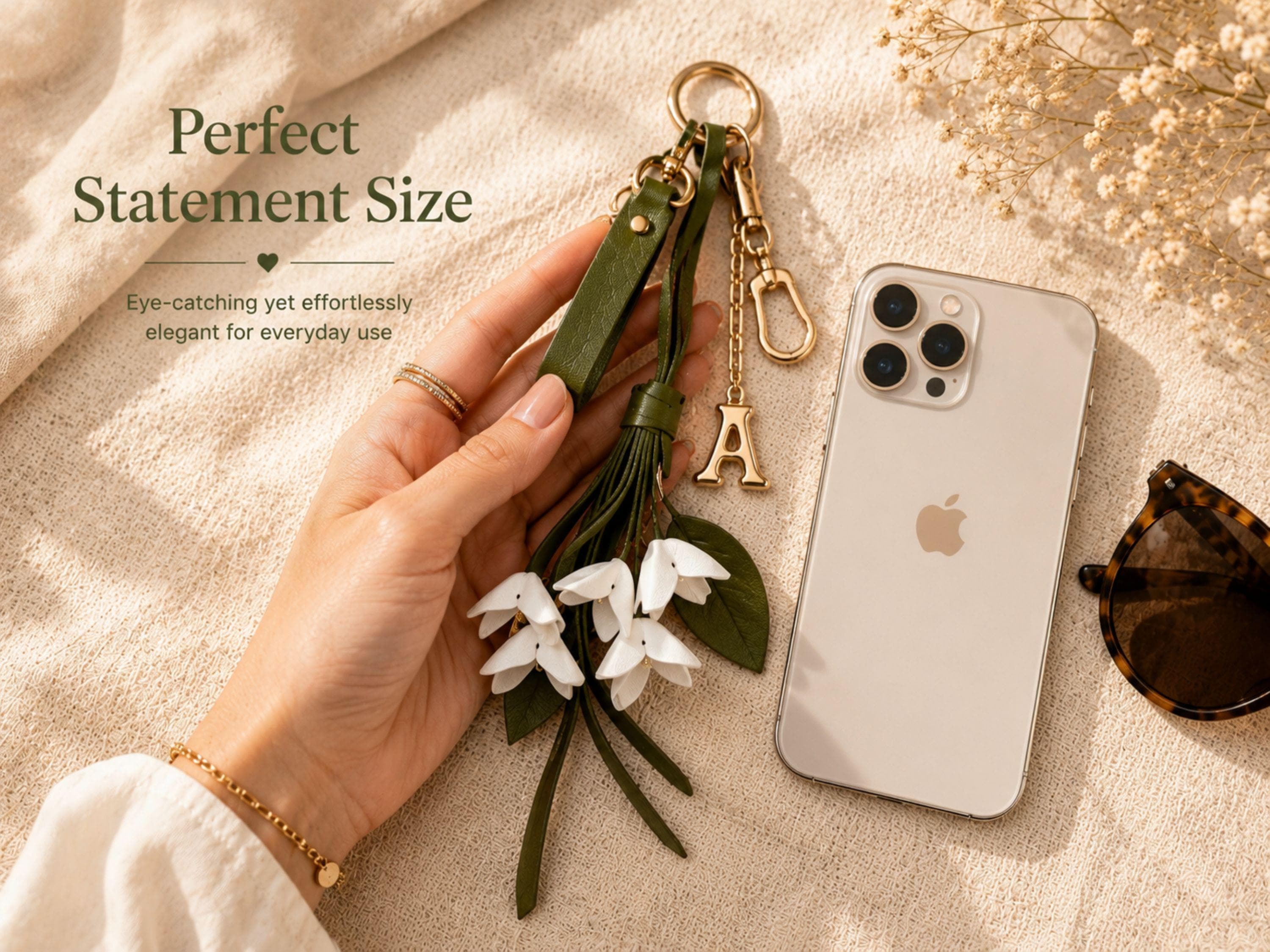 Custom initial bag charm with white folded flowers, green tassel strands, gold hardware and personalized puffy letter charm styled on neutral background. Feminine floral purse accessory for straw tote bags and handbags. Elegant personalized keychain gift for women with quiet luxury aesthetic and summer handbag styling.