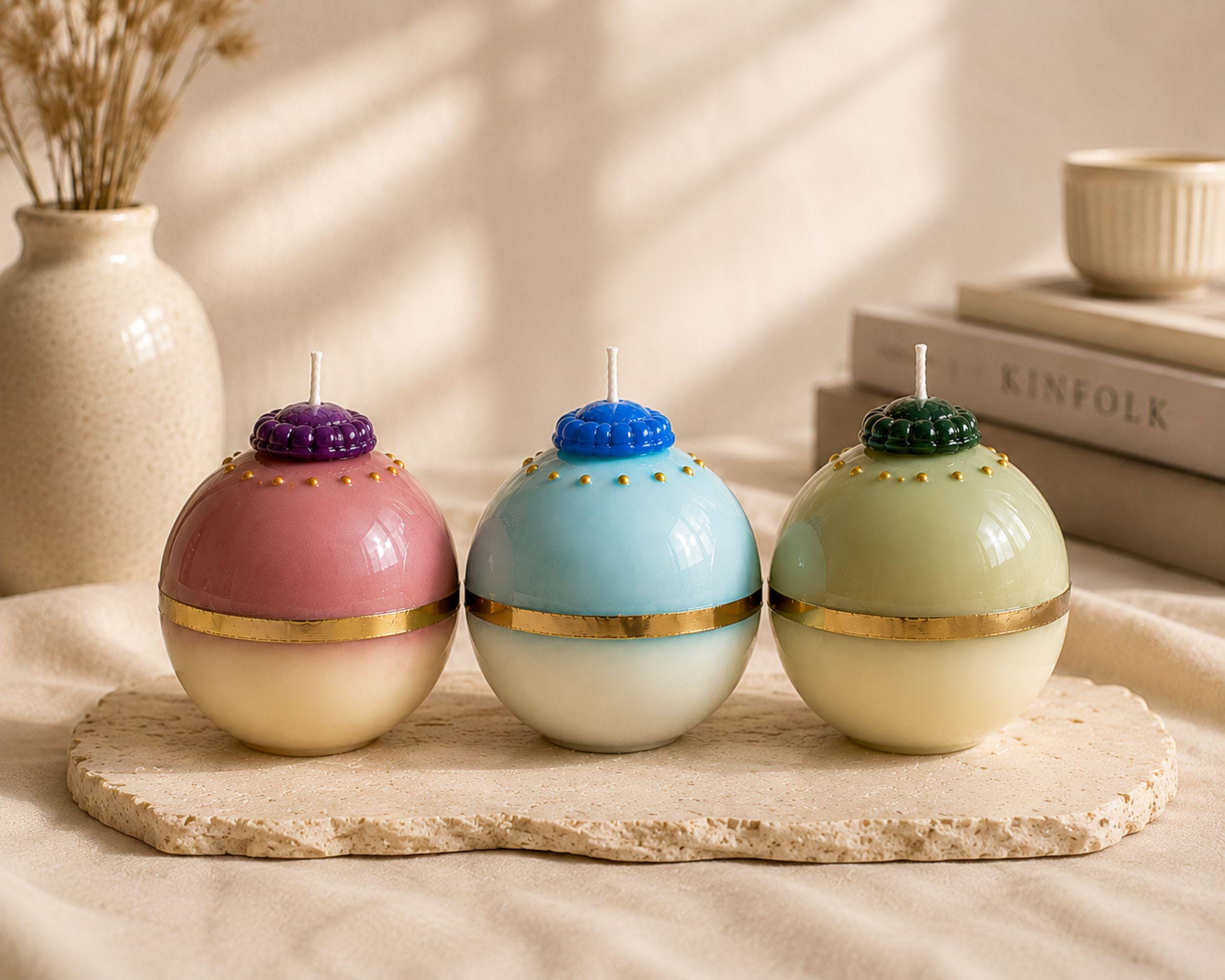 Colorful handmade sphere candles displayed on a neutral stone tray in an elegant interior. Pink and purple, aqua blue, and pale green decorative ball candles with metallic gold details create unique coffee table decor, shelf decor and sculptural home accents.